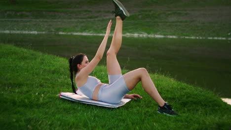 A-woman-does-exercises-for-the-muscles-of-the-press-lying-on-the-grass-in-a-Park-near-the-lake.-Work-with-abs-muscles.-Creating-a-beautiful-body